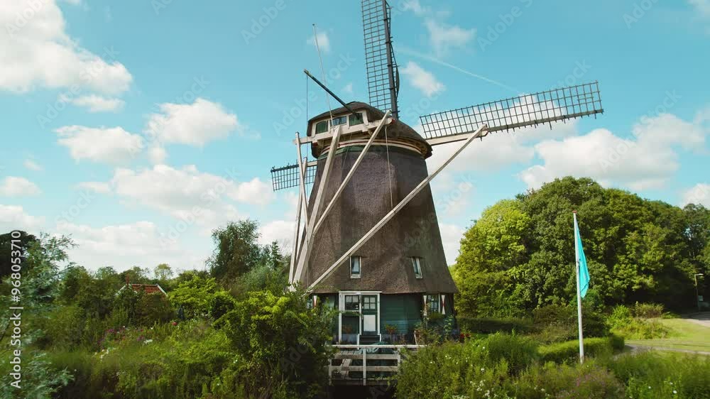 A traditional Dutch windmill stands against a backdrop of blue sky, green fields, and fluffy clouds on a sunny day. A traditional Dutch windmill stands against a backdrop of blue sky, green fields, and fluffy clouds on a sunny day.