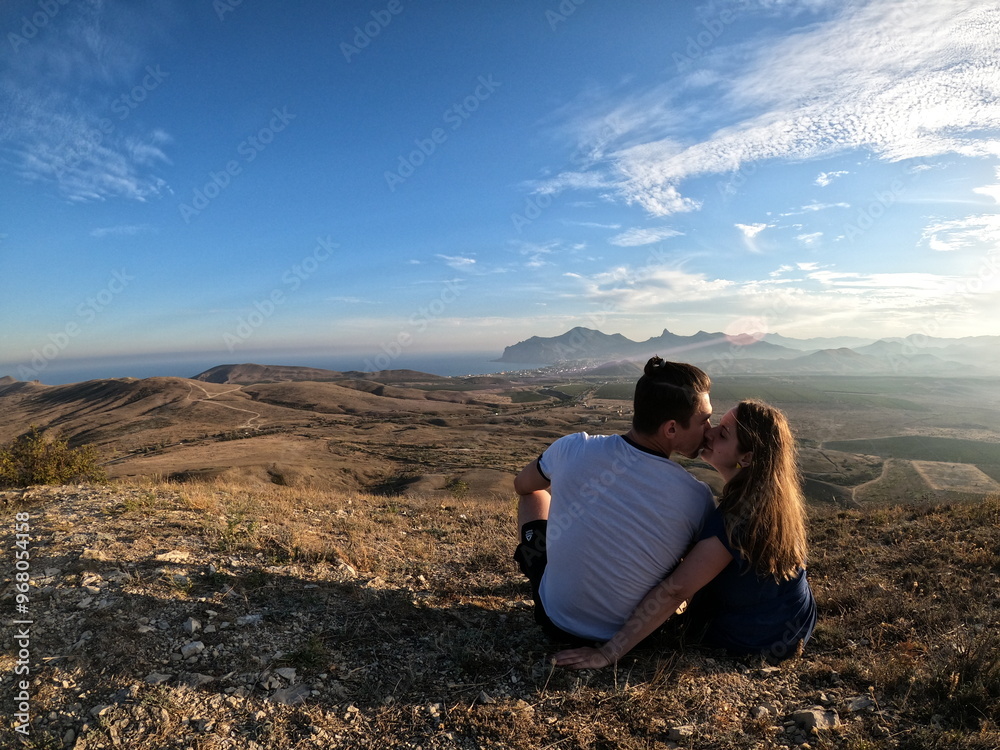 Fototapeta premium Couple sitting on a hilltop overlooking a vast mountainous landscape, sharing a kiss while enjoying the sunset under a partly cloudy sky during a peaceful outdoor moment surrounded by nature