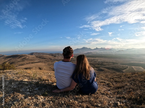 Billede på lærred Couple sitting together on a hilltop, gazing at the vast mountainous landscape u