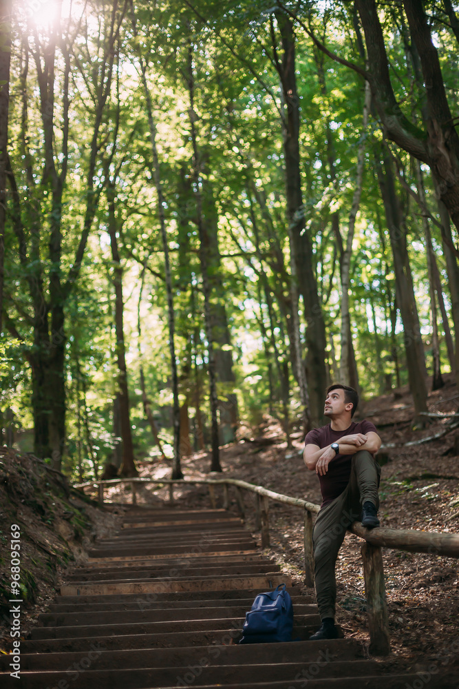 Young man resting on a forest trail staircase, looking up at the sunlight filtering through tall trees, peaceful nature scene in a green forest Young man resting on a forest trail staircase, looking up at the sunlight filtering through tall trees, peaceful nature scene in a green forest