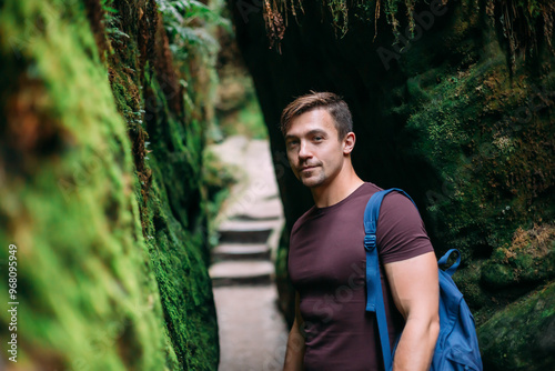 A male tourist with a blue backpack stands near a stone wall covered with bright green moss. A man walks through the Saxon Switzerland National Park in Germany