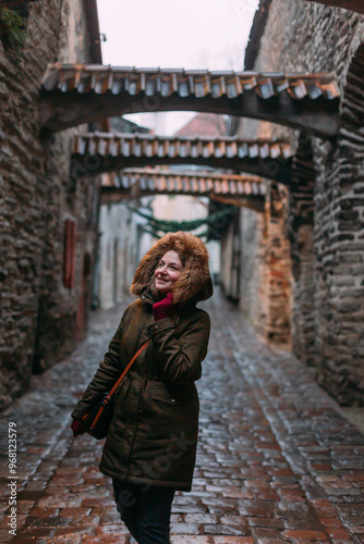 A girl in a marsh-colored winter jacket (khaki) on the street of an old European city. The street is paved with ancient paving stones. The hood is on, there is fur and burgundy gloves