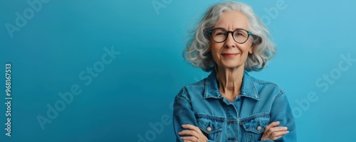 A confident senior woman with a bright smile, standing against a blue background in a casual denim jacket. Free copy space for banner.