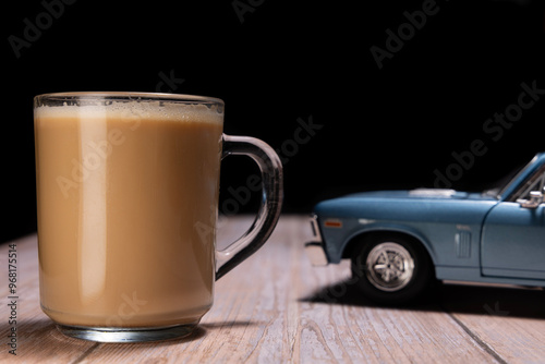 A creative composition featuring a close-up of a frothy coffee in a clear glass mug, with a blue vintage toy car in the background on a wooden surface