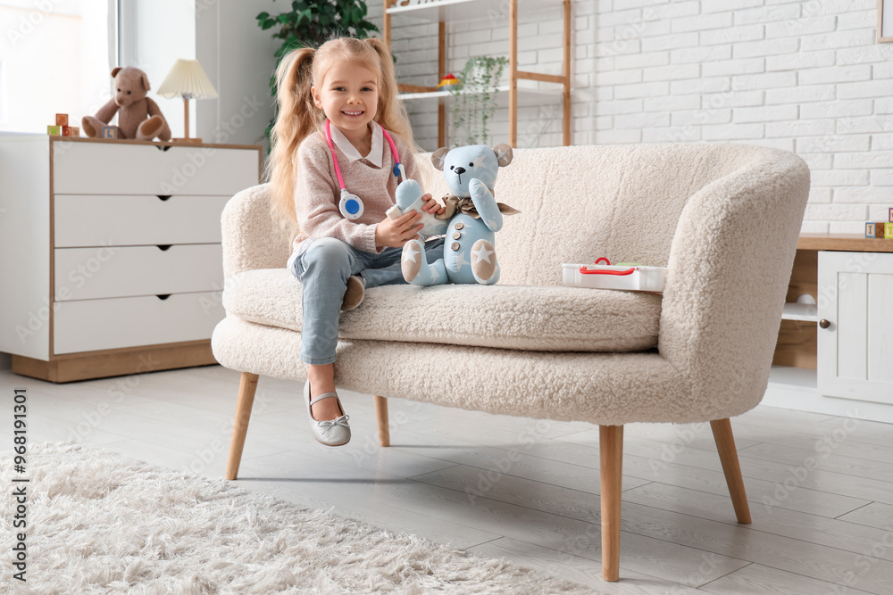 Obraz premium Cute little girl with first aid kit and teddy bear playing on sofa at home