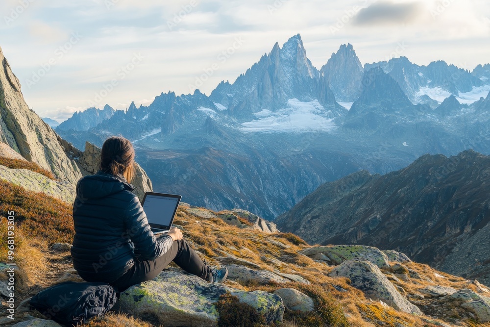 Fototapeta premium An individual sits on a rocky terrain with a stunning view of mountains, working on a laptop surrounded by the awe-inspiring beauty of high-altitude nature.