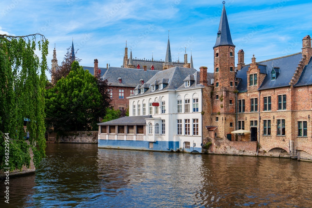 A picturesque view of a canal in Bruges, Belgium. The calm water reflects the charming buildings along the banks, with a clear blue sky and fluffy clouds enhancing the serene atmosphere. A picturesque view of a canal in Bruges, Belgium. The calm water reflects the charming buildings along the banks, with a clear blue sky and fluffy clouds enhancing the serene atmosphere.