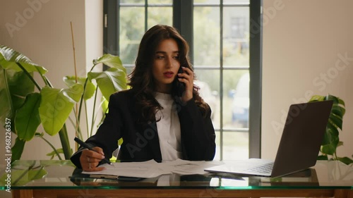 Young Indian woman is sitting at the table at office, talking seriously on the phone and checking something on the computer.