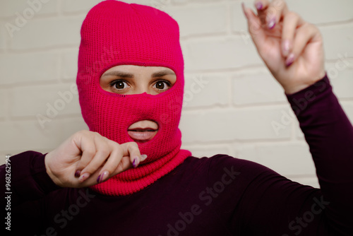 A woman wearing a bright pink balaclava dances energetically, striking unique poses with her hands against a clean, white brick wall background