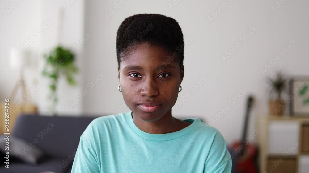 Young serious black woman looking at camera standing in the living room at home. Youth, confidence and African people concept. Young serious black woman looking at camera standing in the living room at home. Youth, confidence and African people concept.