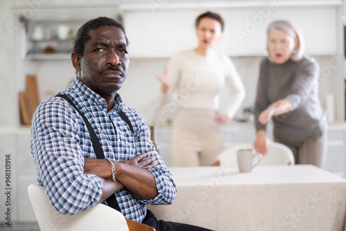 Offended middle-aged man sitting in the kitchen and two women brawling to him angrily standing behind photo