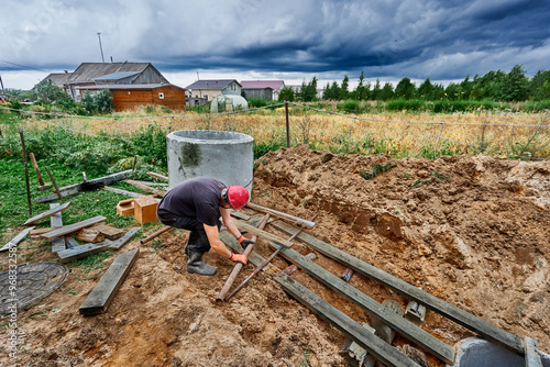 Worker moves concrete ring along wooden platform with steel pipe rollers during installation of septic tank. photo