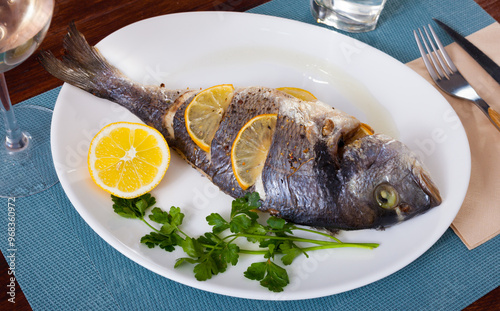 Top view of whole grilled dorada served with lemon and greens at plate, glass of wine photo