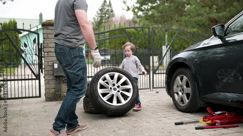 Cute toddler boy helping his father to change car wheels in their backyard. Cute toddler boy helping his father to change car wheels in their backyard.