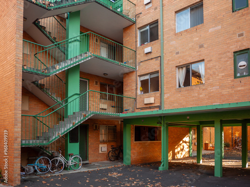 A modest and functional brick apartment building with multiple stories,a walkup staircase on side leading to homes,covered parking area with a few bikes on ground level. North Melbourne,Australia