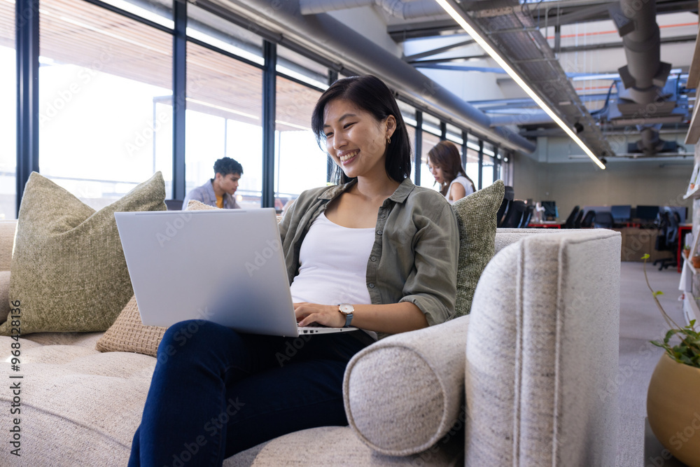 Smiling asian woman using laptop while sitting on couch in modern office Smiling asian woman using laptop while sitting on couch in modern office