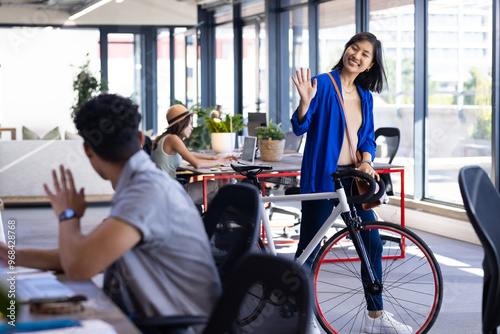 Waving hello, asian woman with bicycle greeting colleague in modern office photo