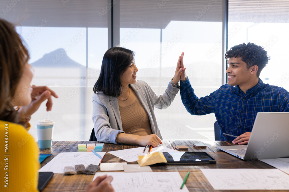 High-fiving, diverse colleagues celebrating success with laptop and design materials in office
