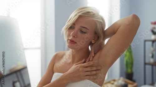 Woman with short blonde hair wrapped in a towel examining her underarm in a spa or beauty salon with light blue interior