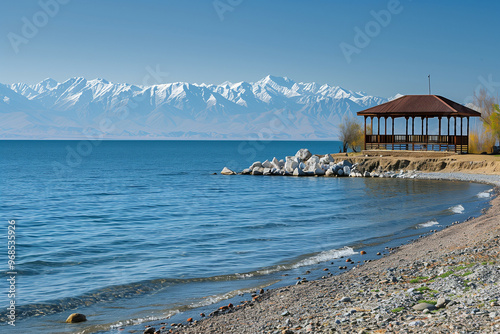 Wallpaper Mural A Serene View of Issyk-Kul Lake in Kyrgyzstan Featuring Snow-Capped Mountains, Tranquil Blue Waters, Pebble Beach, and a Wooden Pavilion Under a Clear Blue Sky with Distant Majestic Peaks Torontodigital.ca