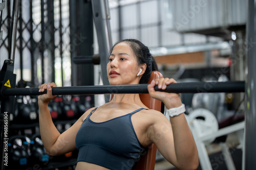 An attractive, sporty Asian woman is lifting a barbell on an incline bench press machine in a gym.