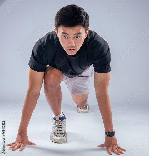 An Asian man in a ready-to-sprint stance, wearing a black t-shirt, gray shorts, and athletic shoes, poised for a workout or race. The setting is a studio with a neutral light blue background photo