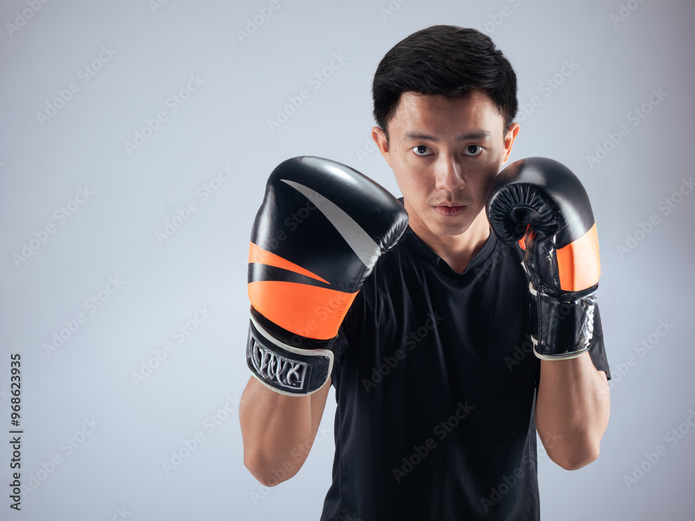 An Asian man in a focused boxing stance, wearing black and orange boxing gloves. He is dressed in a black t-shirt and stands against a white background, ready for action in a fitness studio setting An Asian man in a focused boxing stance, wearing black and orange boxing gloves. He is dressed in a black t-shirt and stands against a white background, ready for action in a fitness studio setting