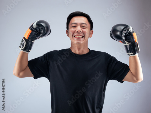 An Asian man wearing black and orange boxing gloves raises his arms triumphantly while smiling in celebration. He is dressed in a black t-shirt, standing against a white background in a studio setting photo