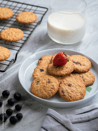 A plate of freshly baked oatmeal raisin cookies garnished with a strawberry, accompanied by a glass of milk and scattered blueberries, set against a light, natural background for a cozy snack scene.