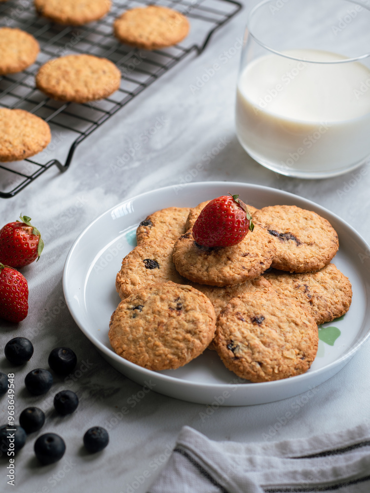 A plate of freshly baked oatmeal raisin cookies garnished with a strawberry, accompanied by a glass of milk and scattered blueberries, set against a light, natural background for a cozy snack scene.