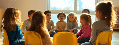 A group of young children of various ethnicities sitting in a circle on yellow chairs in a bright classroom, engaged in conversation and interaction.