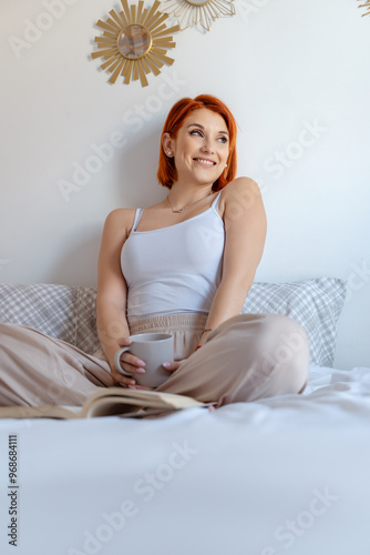 Red-haired woman smiles warmly while holding a coffee cup and sitting on a bed, with an open book nearby. 