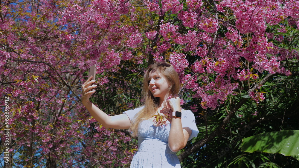 Fototapeta premium Young woman taking selfie in front of beautiful blooming pink cherry blossoms on sunny day, enjoying nature and capturing memories, Connection with Nature and Self-Love.