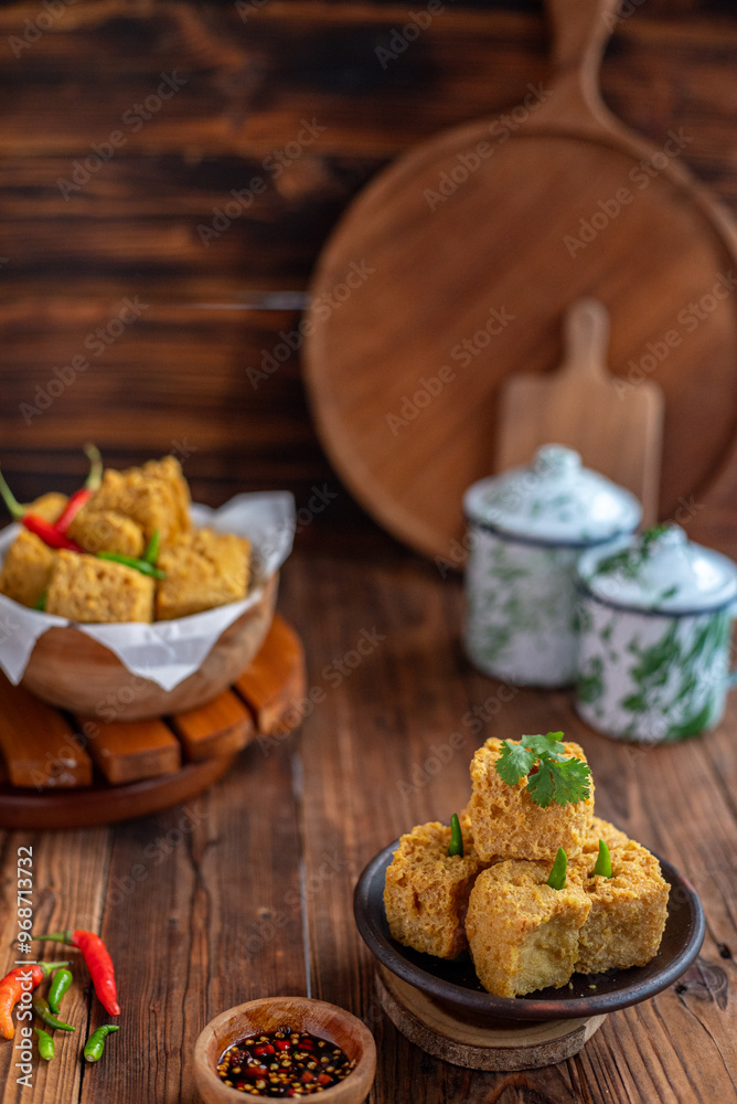 A serving of crispy fried tofu cubes garnished with green chili, presented in a rustic bowl on a wooden table. The background features a blurred bowl of tofu and traditional ceramic containers A serving of crispy fried tofu cubes garnished with green chili, presented in a rustic bowl on a wooden table. The background features a blurred bowl of tofu and traditional ceramic containers