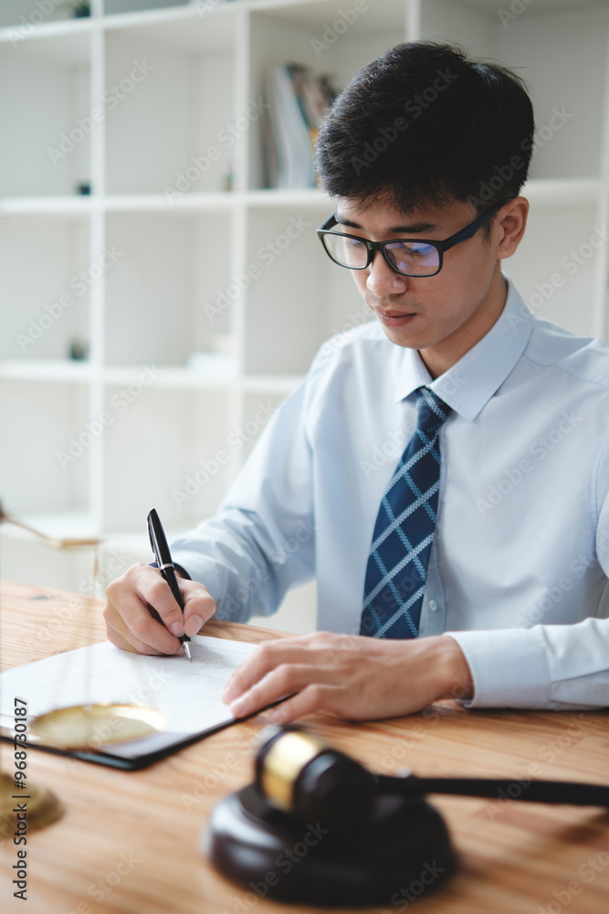 A lawyer in a suit is sitting at his desk, signing legal documents with a gavel on the side.