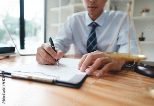 A lawyer in a suit is sitting at his desk, signing legal documents with a gavel on the side.