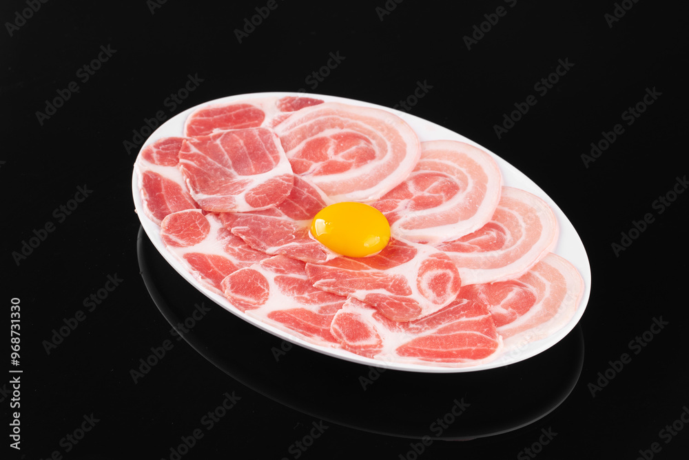 Delicious plate of raw pork slices and an egg yolk on a white dish, ready for cooking, against a black background. Delicious plate of raw pork slices and an egg yolk on a white dish, ready for cooking, against a black background.
