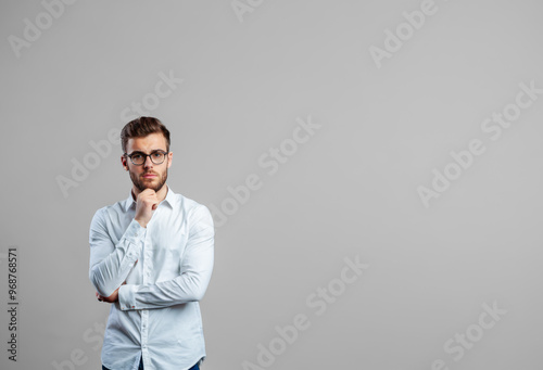 pensive young adult male, neutral backdrop. He wears white shirt, arms crossed, deep in thought. modern business casual style, confident expression