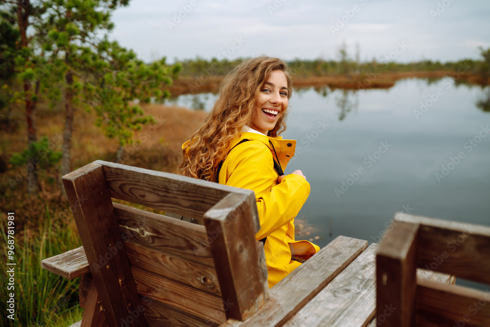 A smiling woman in a yellow raincoat enjoys a walk on a wooden path through a marshy landscape during a cloudy day. Swamps. Travel concept, exploring. A smiling woman in a yellow raincoat enjoys a walk on a wooden path through a marshy landscape during a cloudy day. Swamps. Travel concept, exploring.