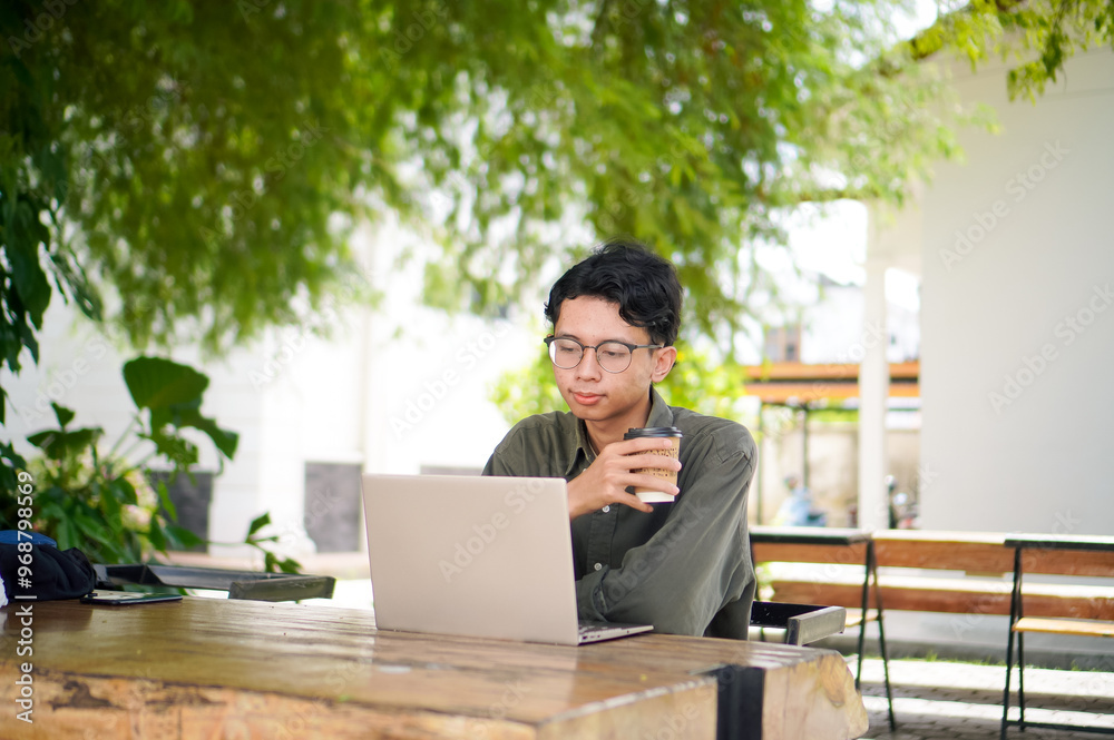 Young asian man thinking while looking to his laptop. Man working on his laptop at the outdoor cafe in the morning Young asian man thinking while looking to his laptop. Man working on his laptop at the outdoor cafe in the morning