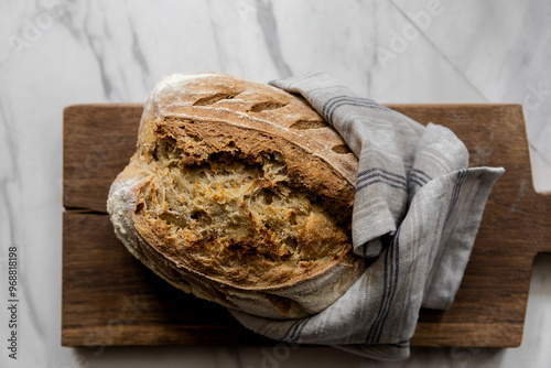 Sourdough bread wrapped in a grey linen towel arranged on a wooden cutting board that is placed on a white marble kitchen counter top  