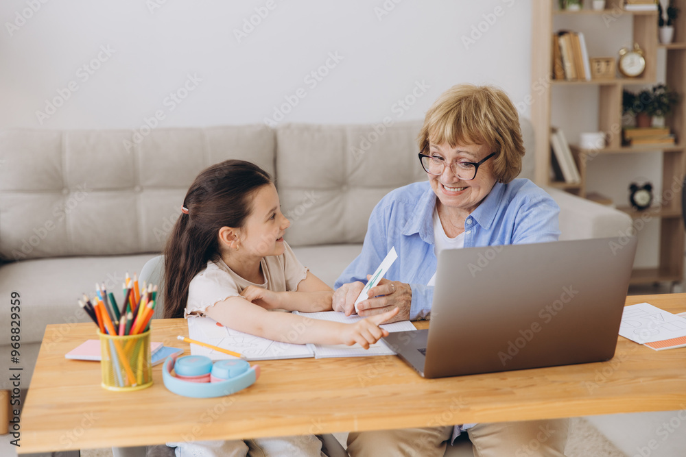 Happy grandma helping adorable preschool granddaughter school kid with homework task. Positive school pupil girl and granny reading learning book at laptop together, enjoying education at home Happy grandma helping adorable preschool granddaughter school kid with homework task. Positive school pupil girl and granny reading learning book at laptop together, enjoying education at home