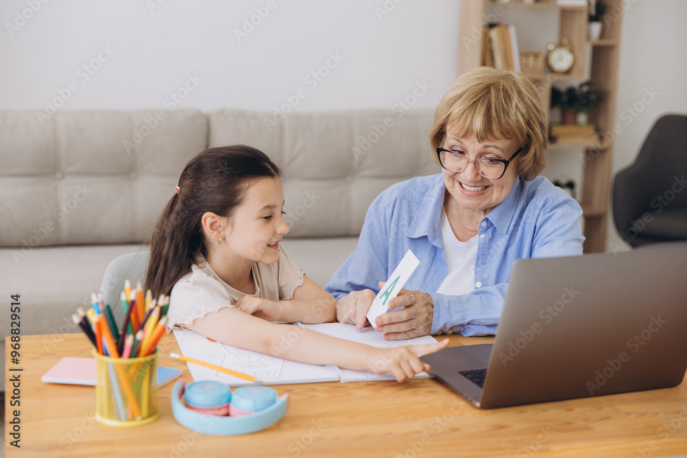 Happy grandma helping adorable preschool granddaughter school kid with homework task. Positive school pupil girl and granny reading learning book at laptop together, enjoying education at home Happy grandma helping adorable preschool granddaughter school kid with homework task. Positive school pupil girl and granny reading learning book at laptop together, enjoying education at home