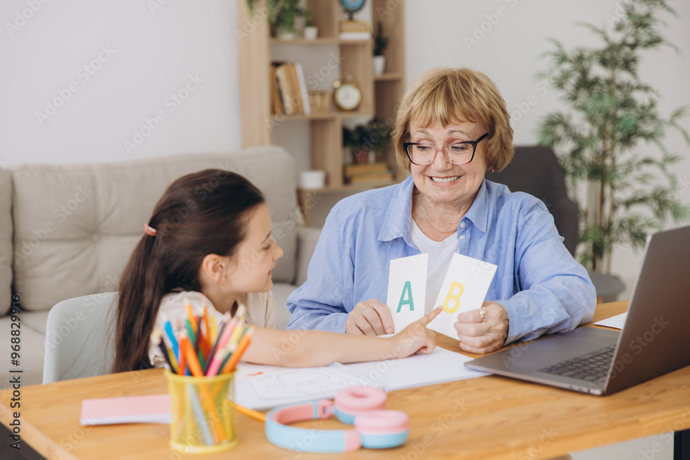 Happy grandma helping adorable preschool granddaughter school kid with homework task. Positive school pupil girl and granny reading learning book at laptop together, enjoying education at home Happy grandma helping adorable preschool granddaughter school kid with homework task. Positive school pupil girl and granny reading learning book at laptop together, enjoying education at home