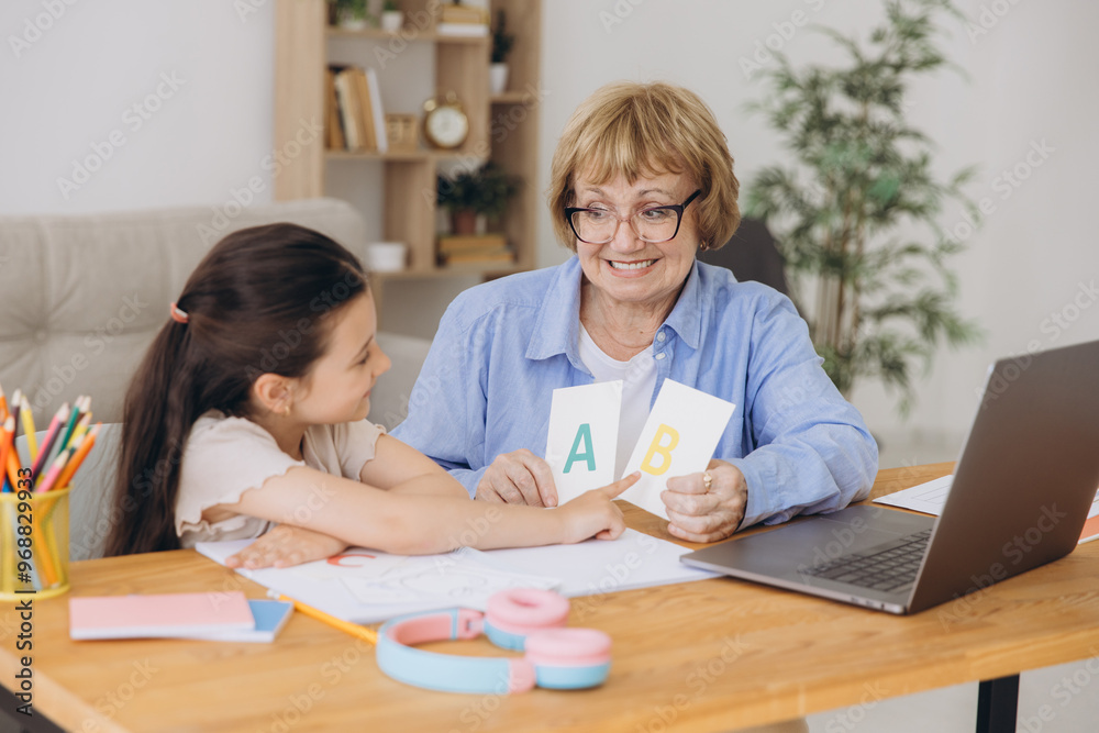 Happy grandma helping adorable preschool granddaughter school kid with homework task. Positive school pupil girl and granny reading learning book at laptop together, enjoying education at home Happy grandma helping adorable preschool granddaughter school kid with homework task. Positive school pupil girl and granny reading learning book at laptop together, enjoying education at home