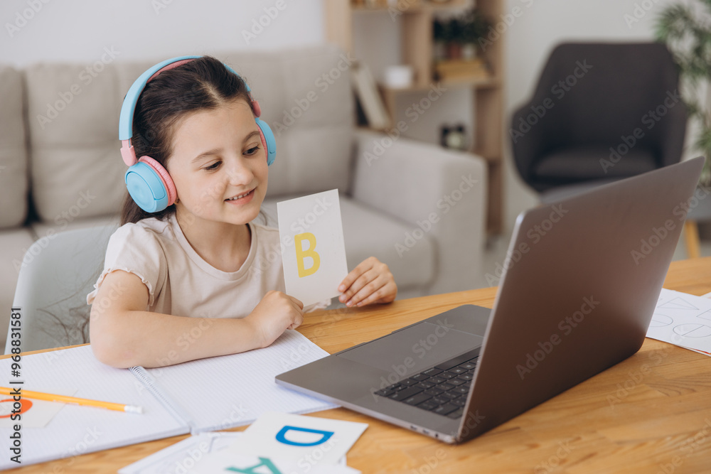 Smiling small girl in headphones watch video lesson on computer with alphabet, happy little child in earphones have online web class using laptop at home, homeschooling concept Smiling small girl in headphones watch video lesson on computer with alphabet, happy little child in earphones have online web class using laptop at home, homeschooling concept