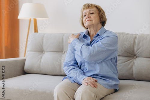 Senior woman sitting on sofa with shoulder pain. Elderly woman is enduring awful ache. Old woman suffering from shoulder injury, health problem concept