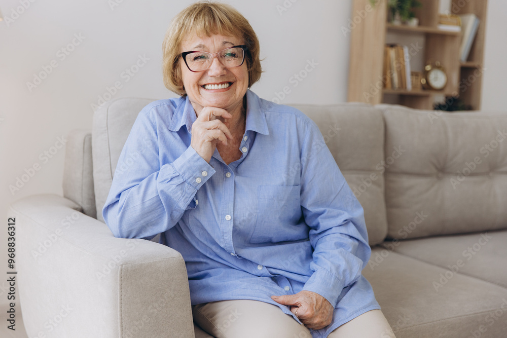 Smiling senior blonde haired woman looking at camera, happy old lady in glasses posing at home indoor, positive single senior retired female sitting on sofa in living room headshot portrait Smiling senior blonde haired woman looking at camera, happy old lady in glasses posing at home indoor, positive single senior retired female sitting on sofa in living room headshot portrait