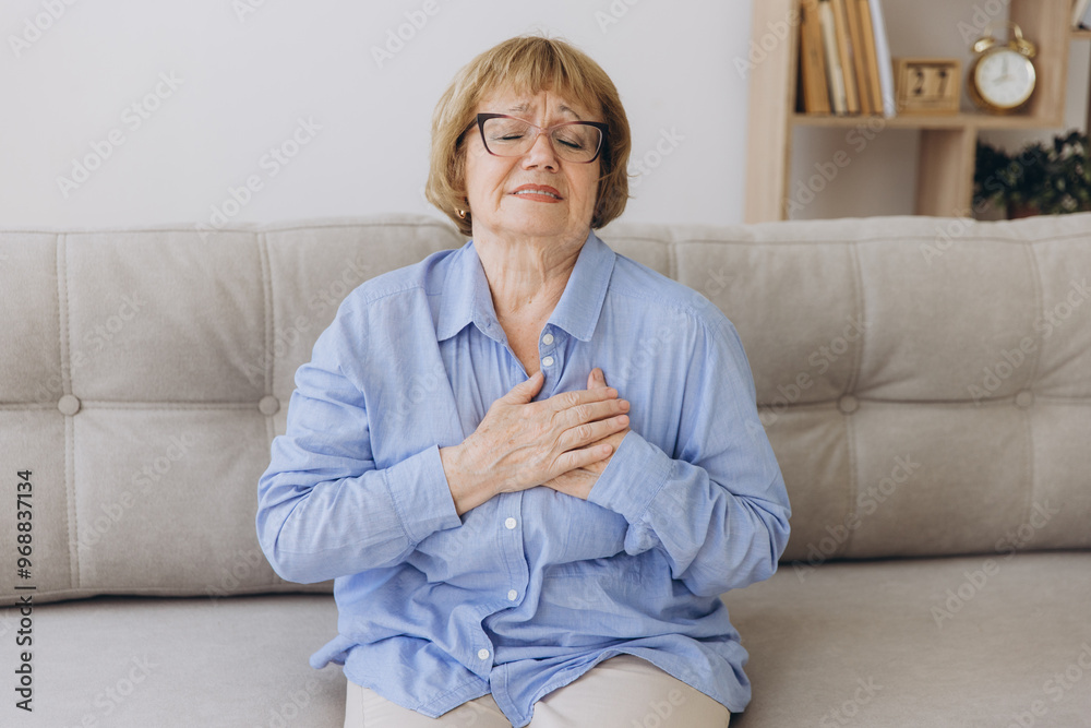 Elderly senior woman feeling heart pain and holding her chest while sitting on the sofa in her living room. Elderly senior woman feeling heart pain and holding her chest while sitting on the sofa in her living room.