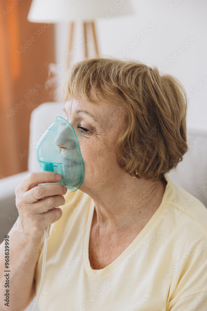 Sick senior woman has a viral infection or flu, labored breathing, asthma. An elderly female is using inhaler, nebulizer for self treatment sitting alone on the sofa at home Sick senior woman has a viral infection or flu, labored breathing, asthma. An elderly female is using inhaler, nebulizer for self treatment sitting alone on the sofa at home
